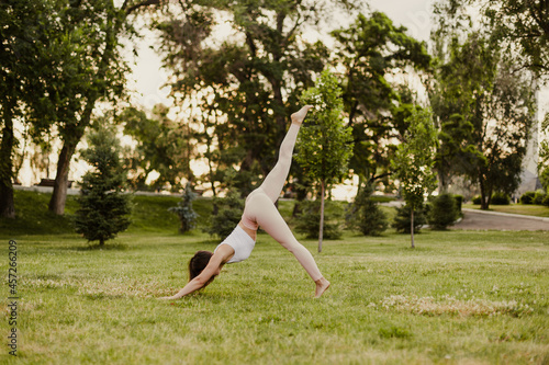 full-length young woman practices yoga in Downward-Facing Dog pose with one leg raised up in nature in park, concept of restoring and slowing down life. 