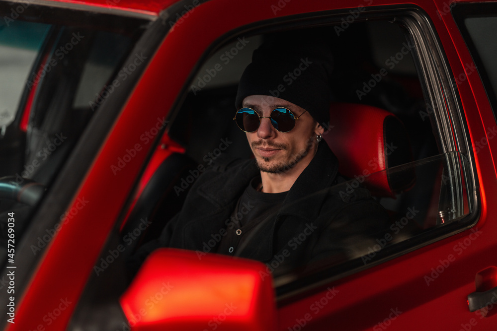 Handsome cool man with hat and black jacket with sunglasses drives a red car. Travel by the car Handsome cool man with hat and black jacket with sunglasses drives a red car. Travel by the car