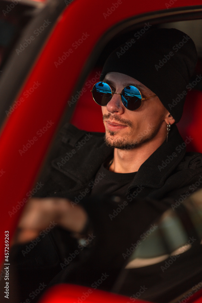stylish handsome man with sunglasses in hat and coat sits behind the wheel of a red old car stylish handsome man with sunglasses in hat and coat sits behind the wheel of a red old car