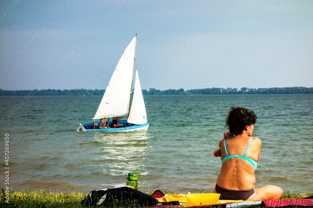 Fototapeta premium A young woman sits in a swimsuit on the beach by the lake and sunbathes, in the background is a sailing yacht with a crew