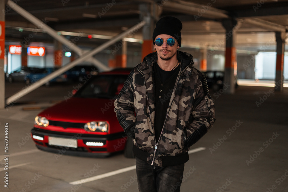 Stylish man hipster with sunglasses and black hat with fashion military winter jacket walks on the street near a red car at parking lot