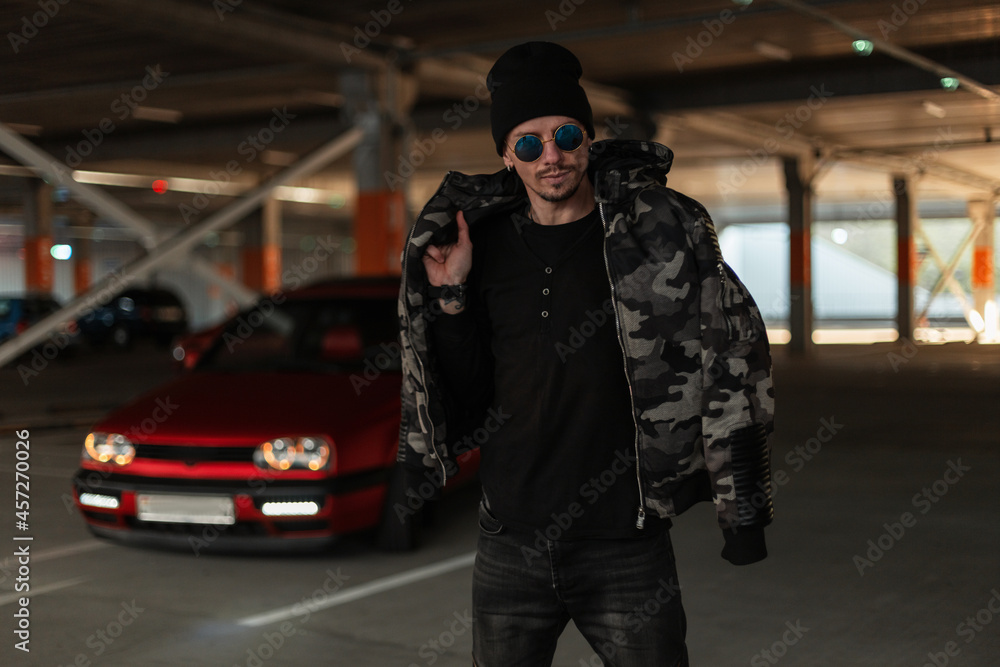Handsome fashionable man model with cool sunglasses in a stylish camouflage winter jacket, pullover and hat walks on the street near a red car and a parking lot Handsome fashionable man model with cool sunglasses in a stylish camouflage winter jacket, pullover and hat walks on the street near a red car and a parking lot