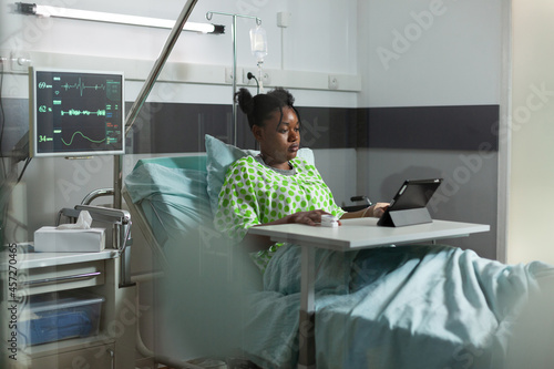 Young african american woman using tablet in hospital ward bed. Sick patient with online technology recovering from disease at clinic with medical equipment. Person web surfing photo