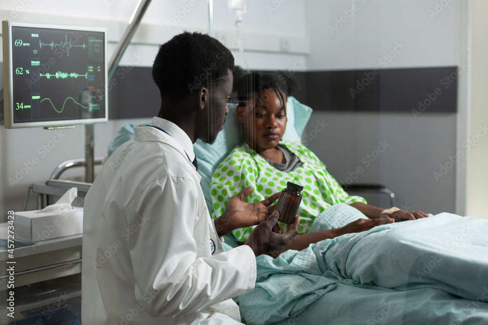 Specialist explaining treatment to ill patient in hospital ward at clinic. African american doctor showing bottle of drugs, pills on prescription for healthcare medicine and immunity Specialist explaining treatment to ill patient in hospital ward at clinic. African american doctor showing bottle of drugs, pills on prescription for healthcare medicine and immunity