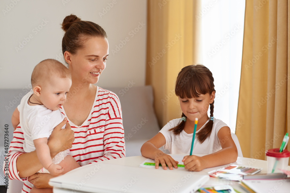 Smiling mother with new born baby in hands and elder daughter sitting at table and doing homework, dark haired girl in white t shirt writing in exercises book or drawing. Smiling mother with new born baby in hands and elder daughter sitting at table and doing homework, dark haired girl in white t shirt writing in exercises book or drawing.