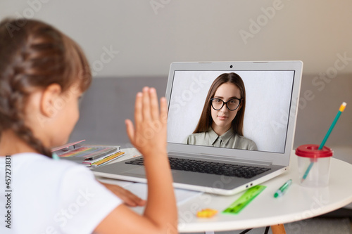 Indoor shot of dark haired girl with pigtails wearing white t shirt  posing backwards  having online lesson with teacher  looking at display and waving hand  online distant education.