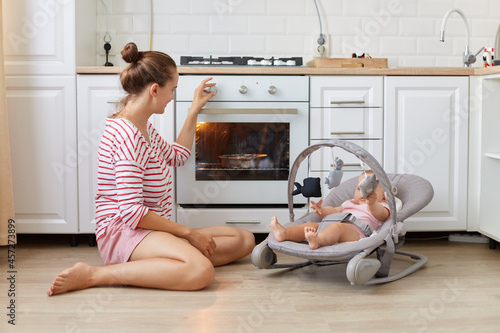 Fotografi Mother baking a pie with her newborn daughter lying in rocking chair, young woman wearing casual style shirt and kid in bouncer cook in a white kitchen, baking pastry, making dinner