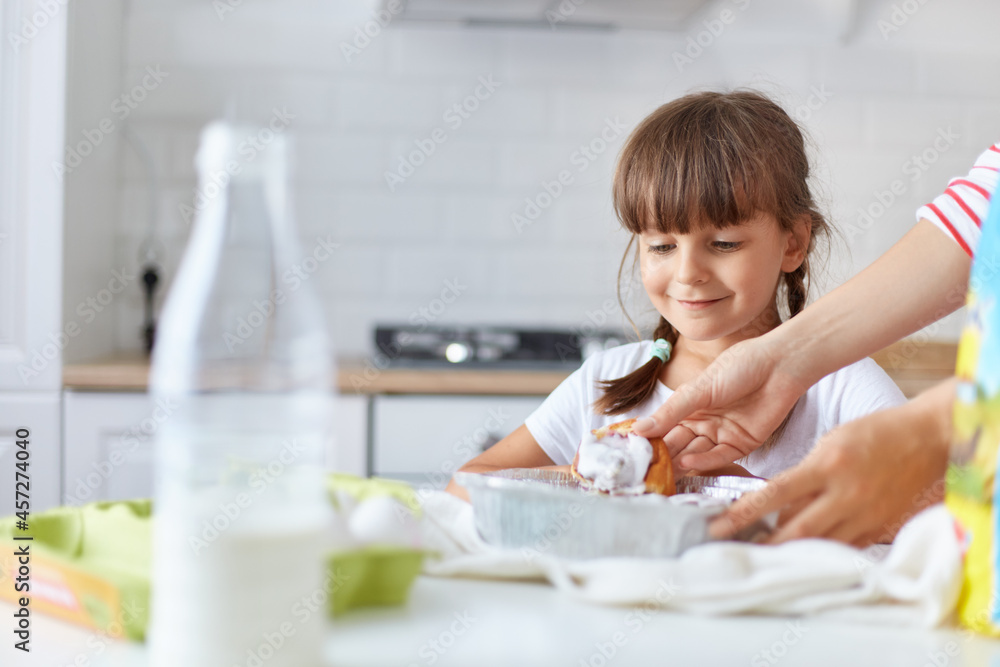 Positive smiling female child with two pigtails standing near table, waiting her mom giving her hot tasty baking, kid girl wearing white t shirt posing in kitchen.