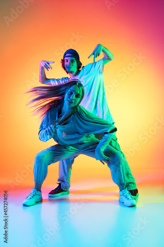 Young hip-hop dancers, stylish emotive girl and boy in action and motion in casual sports youth clothes on gradient multi colored background at dance hall in neon light. © master1305