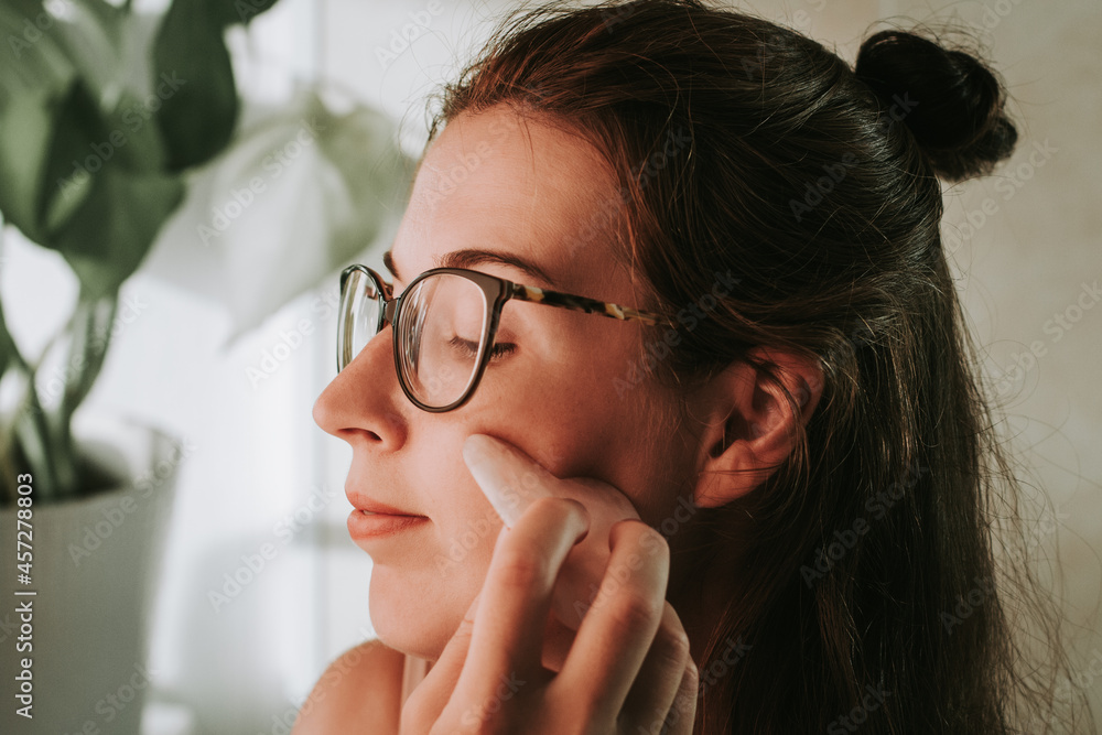 Woman in eyeglasses doing face lifting massage with gua sha in front of a mirror Woman in eyeglasses doing face lifting massage with gua sha in front of a mirror