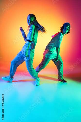 Two dancers, young girl and boy dancing hip-hop in casual sports youth clothes on gradient multi colored background at dance hall in neon light.