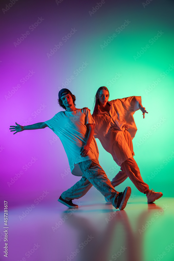 Stylish young hip-hop dancers, emotive girl and boy in action and motion in casual sports youth clothes on gradient multi colored background at dance hall in neon light.