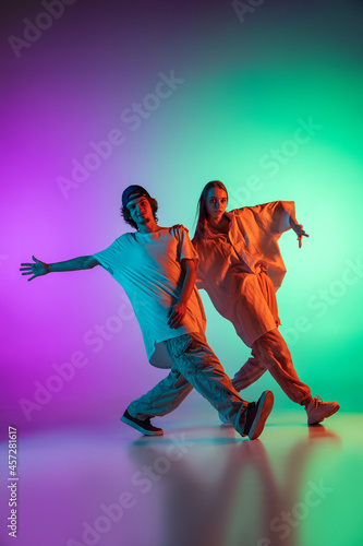 Stylish young hip-hop dancers, emotive girl and boy in action and motion in casual sports youth clothes on gradient multi colored background at dance hall in neon light.