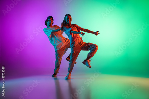 Young hip-hop dancers, stylish emotive girl and boy in action and motion in casual sports youth clothes on gradient multi colored background at dance hall in neon light. © master1305