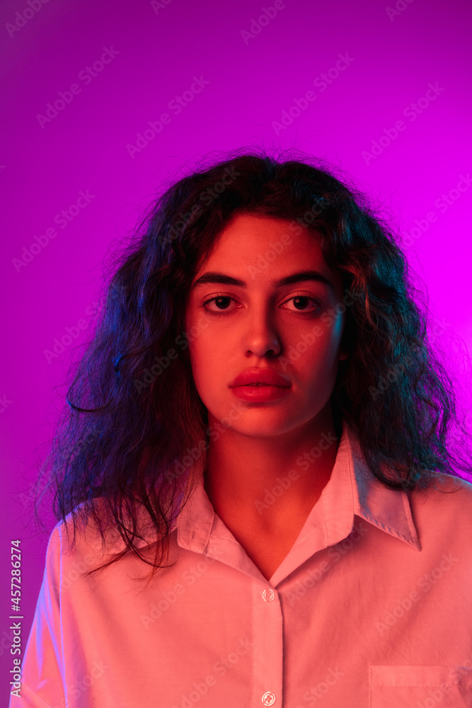 Charming young woman looking at camera isolated on purple, lilac color studio background in neon light. Concept of emotions, expressions Charming young woman looking at camera isolated on purple, lilac color studio background in neon light. Concept of emotions, expressions