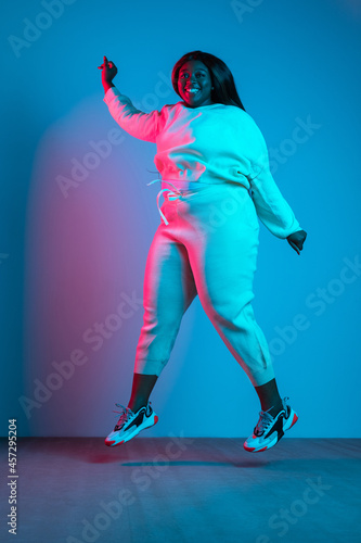 Full-length portrait of African beautiful woman with long straight hair jumping isolated on blue studio background in neon light. Concept of human emotions