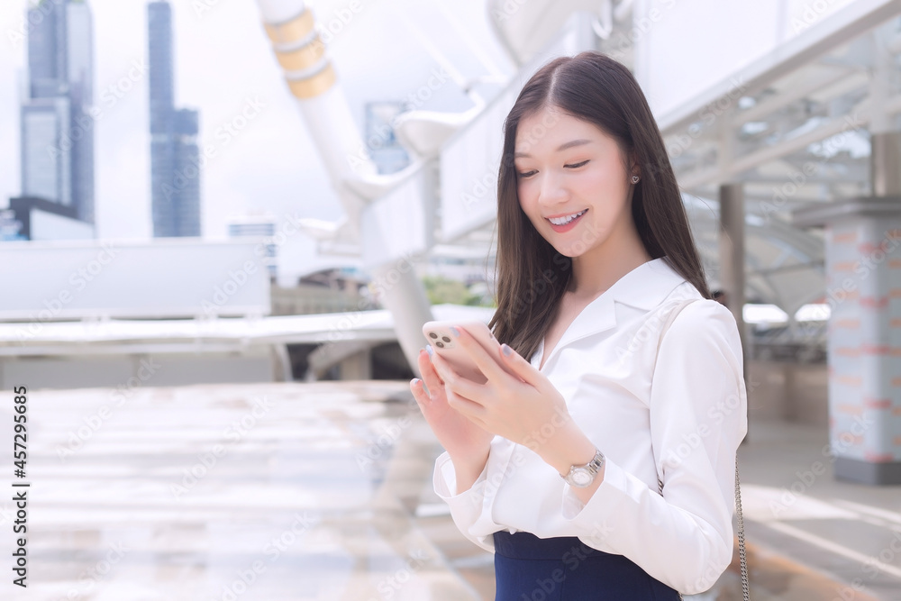 Young Asian Business woman who smiles is playing and looking at smartphone in her hands while she is going to office in business background in the city. Young Asian Business woman who smiles is playing and looking at smartphone in her hands while she is going to office in business background in the city.