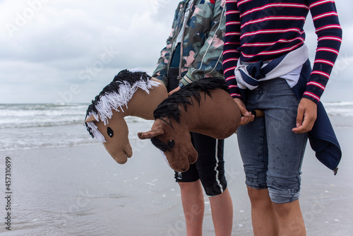 two young girls with their hobbyhorses standing on the beach with the sea in the background