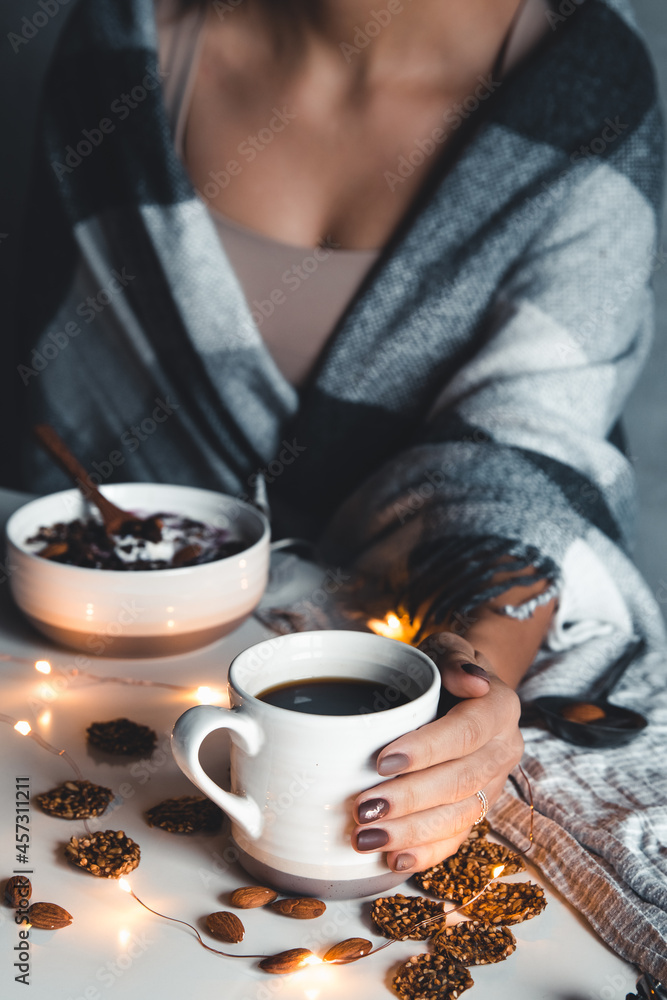 The woman is wrapped in a blanket and holds a cup of coffee in her hands. Winter comfort, garlands, Christmas mood. Stylish manicure, nail art. Healthy food, smoothies on a plate, granola. The woman is wrapped in a blanket and holds a cup of coffee in her hands. Winter comfort, garlands, Christmas mood. Stylish manicure, nail art. Healthy food, smoothies on a plate, granola.