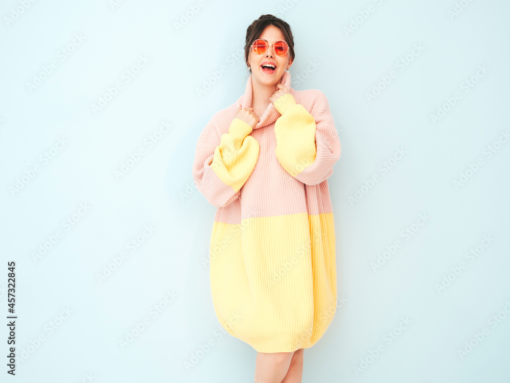 Young beautiful smiling female in trendy summer colorful sweater. Sexy carefree woman posing near light blue wall in studio. Positive model having fun indoors in sunglasses Young beautiful smiling female in trendy summer colorful sweater. Sexy carefree woman posing near light blue wall in studio. Positive model having fun indoors in sunglasses