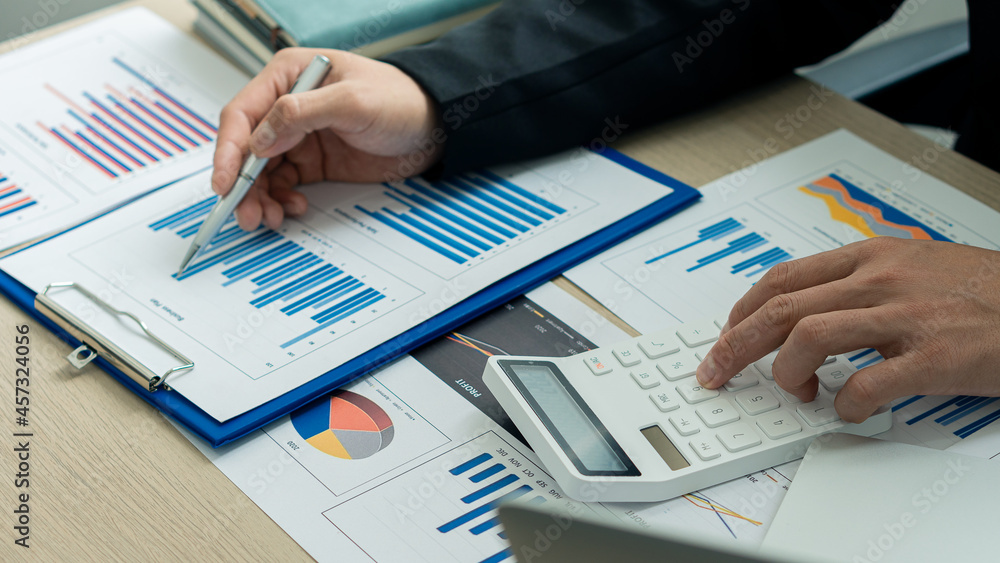 Hands of money making and calculating on desk about home office expenses. An accountant uses a calculator and computer holding a pen pointing to a graph document. Hands of money making and calculating on desk about home office expenses. An accountant uses a calculator and computer holding a pen pointing to a graph document.