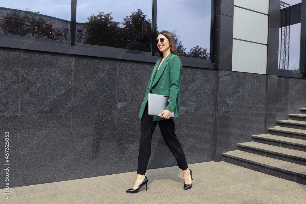 A young woman in a business suit with a laptop is walking near an office building