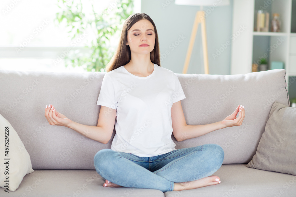 Portrait of attractive calm focused girl sitting on divan in lotus pose meditating resting at home indoors