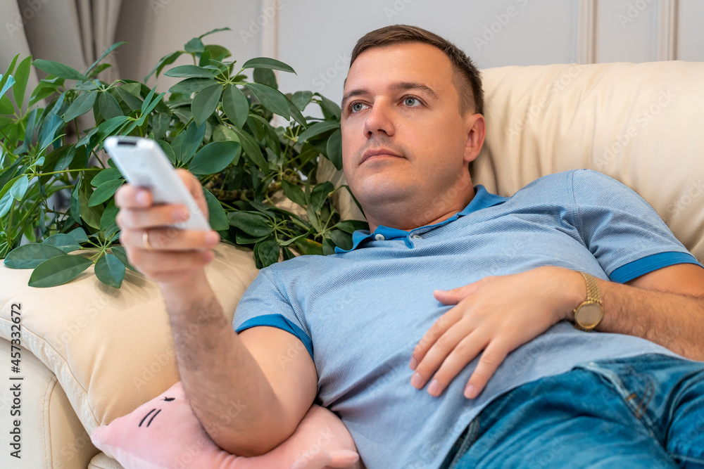 Bored millennial man is watches TV, holds remote control to switch channels. Guy is bored with what he sees on TV screen. Handsome man lies on sofa in living room at home.