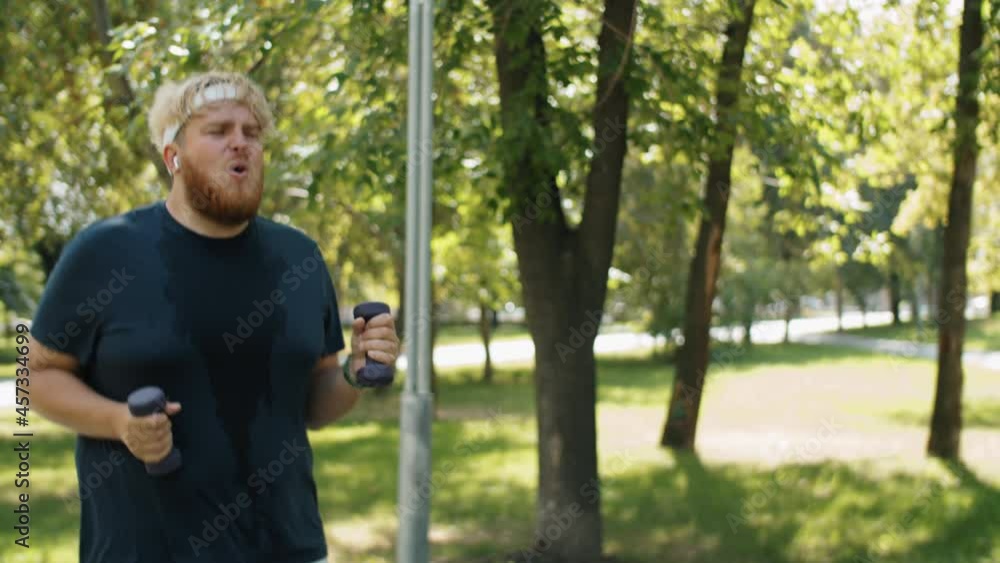 Young overweight man jogging and then doing air punches with dumbbells during fat burning workout in park