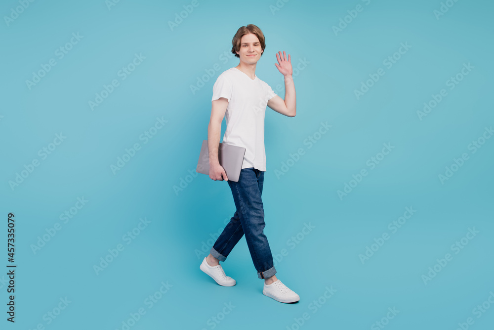 Full body photo of cheerful guy walking wave hand hello carrying laptop isolated on blue background Full body photo of cheerful guy walking wave hand hello carrying laptop isolated on blue background