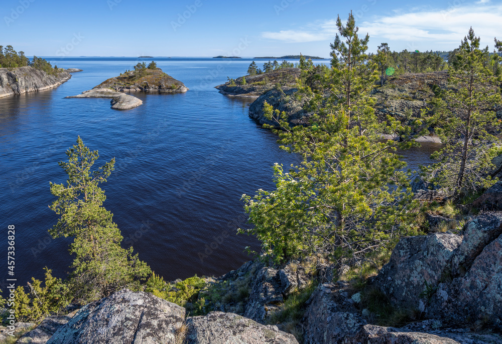 Fototapeta premium Landscape with a forest on stones over the lake. Sunny day at the lake. Reflection of the sky in the water. Pines on stones. The nature of the north.