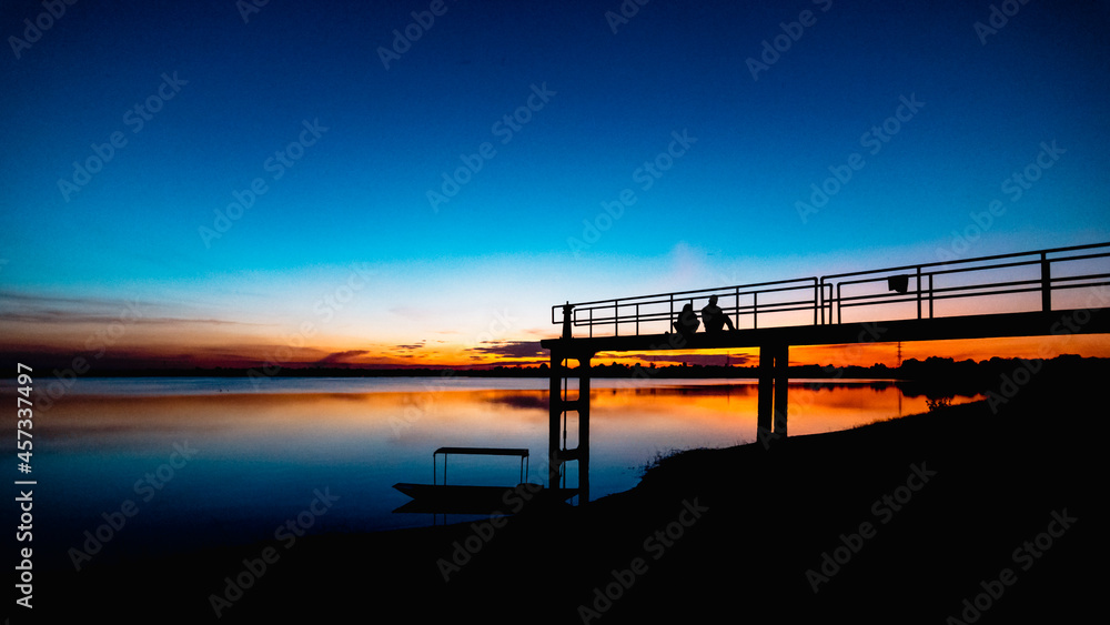 Two silhouette people on the bridge with a sunset background and a boat