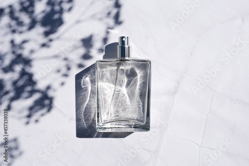 Transparent bottle of perfume with spray on white marble surface. Clear glass without lid. Trending concept in natural materials with plant shadow. Luxury presentation. photo