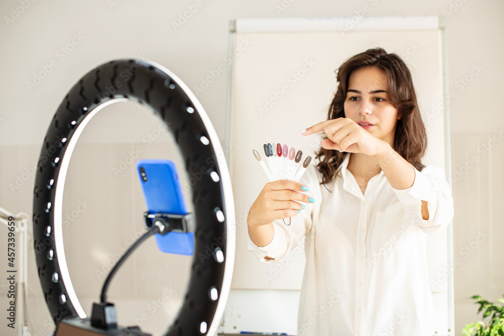 Young woman manicurist master blogger recording video using ring lamp and smartphone, talking and gesturing at camera, explain something. Online Master class, training courses, lesson via internet Young woman manicurist master blogger recording video using ring lamp and smartphone, talking and gesturing at camera, explain something. Online Master class, training courses, lesson via internet