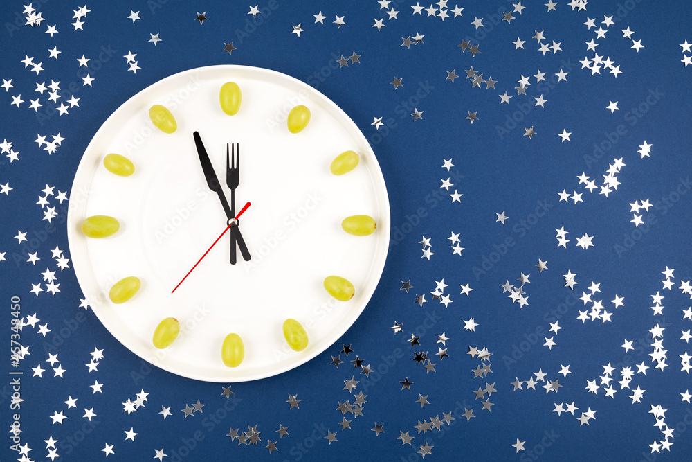 Clock made of grapes on blue background with sequins. Latin American and Spanish New Year tradition to eat twelve 12 grapes for good luck at midnight. Christmas composition. Flat lay, top view Clock made of grapes on blue background with sequins. Latin American and Spanish New Year tradition to eat twelve 12 grapes for good luck at midnight. Christmas composition. Flat lay, top view