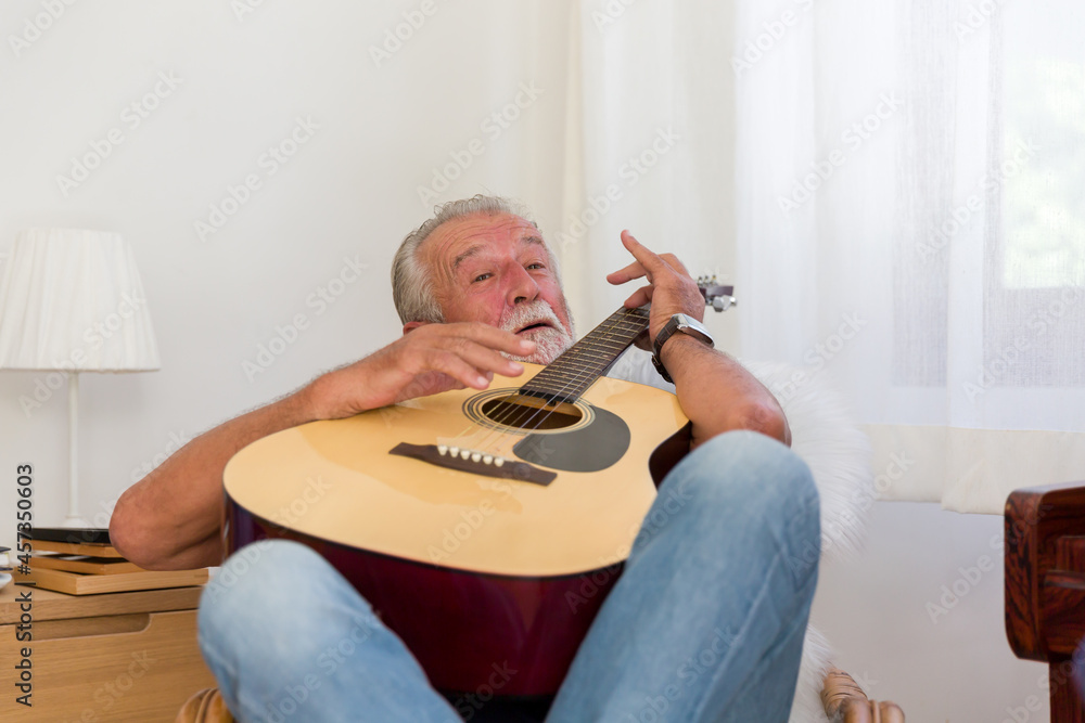 Cheerful senior man playing guitar while sitting on sofa at home. Elderly man learning guitar on holiday. people, quarantine, holiday concept
