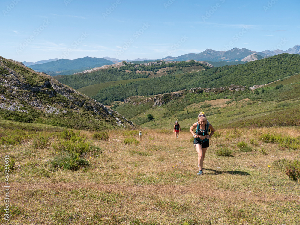 blonde woman hiker going up a green hill surrounded by a beautiful landscape with the mountains behind