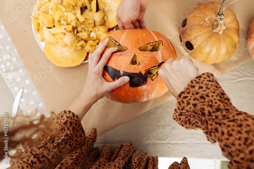 Close up of young woman carving Jack O Lattern from ripe orange pumpkin with knife on her wooden kitchen table. Female preparing all hallows eve Halloween party decorations. Background, copy space. photo