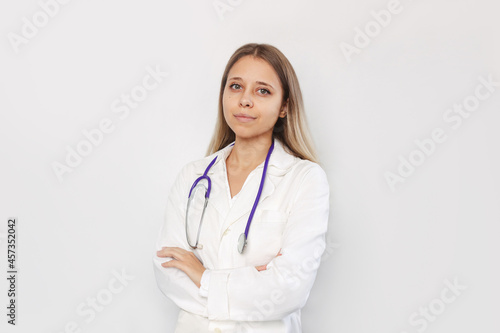 A young blonde female doctor in a white coat with a stethoscope around her neck stands with her hands crossed on her stomach isolated on a white background