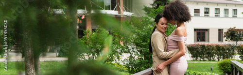 Smiling woman in suit hugging african american girlfriend in dress in park  banner.