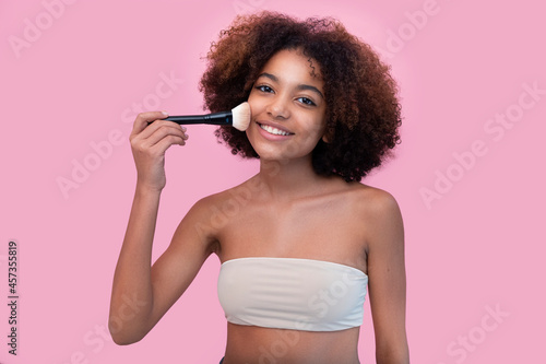 Beauty portrait of a young dark-skinned beautiful woman with curly hair smiling with her eyes closed and stroking her cheek with a makeup brush on a pink background