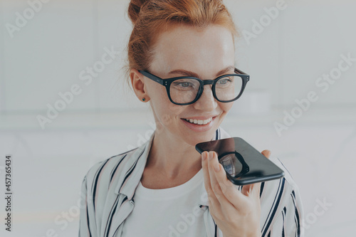 Fotografija Redhead woman in transparent glasses speaks to virtual assistant holds mobile ph