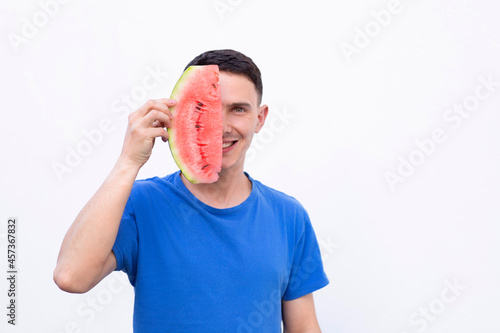 The guy covers half of his face with a slice of watermelon. A guy in a blue T-shirt with a watermelon in his hand. Cheerful young man.
