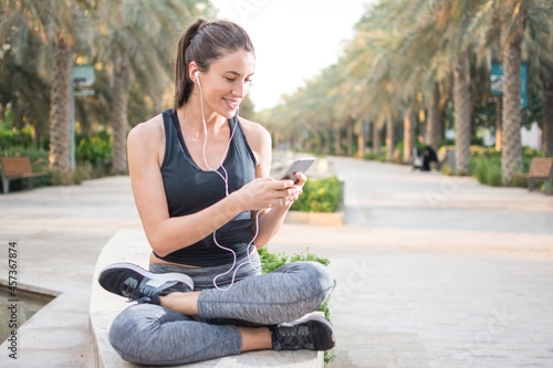 Smiling young woman in running attire sitting outdoors and listening to music from mobile phone.