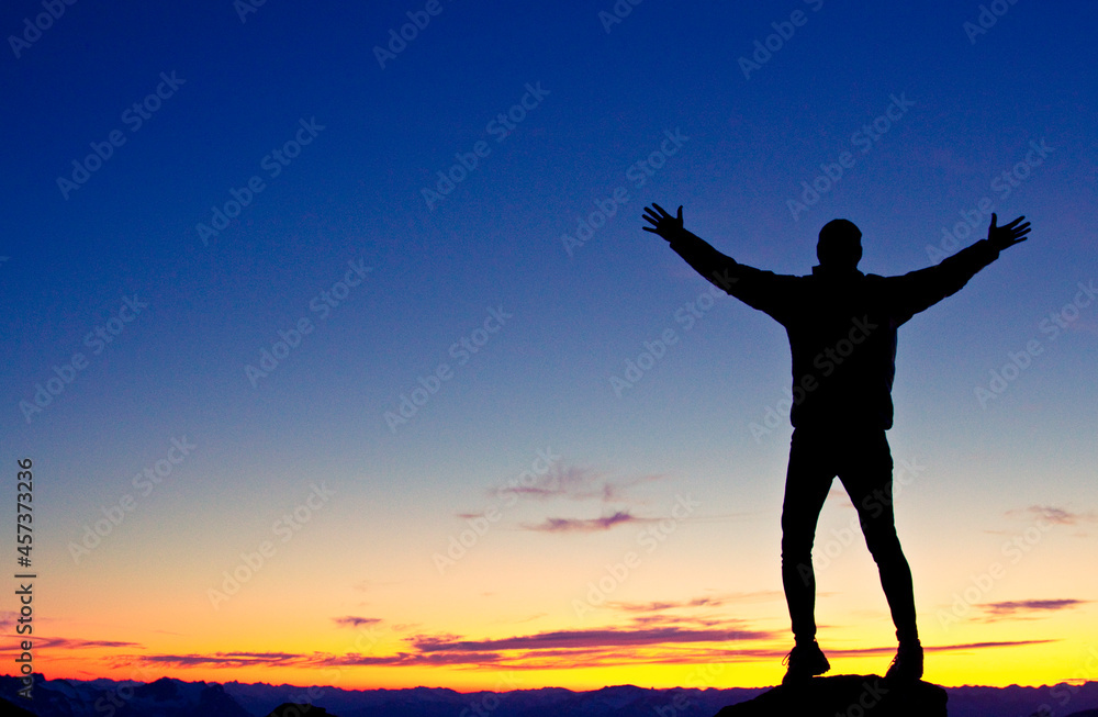 Silhouetted hiker standing on mountain summit with arms raised Silhouetted hiker standing on mountain summit with arms raised