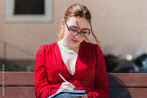 Young red-haired woman in a red blouse sitting on a bench while writing something in her notebook. photo