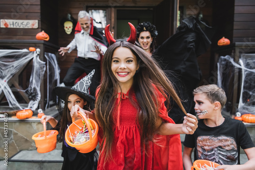 happy girl in devil halloween costume holding bucket of sweets while running out cottage with family