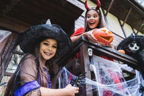 low angle view of cheerful sisters in halloween costumes playing on porch with decoration