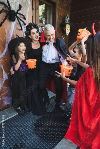 happy family in creepy costumes holding buckets with sweets on porch with halloween decoration