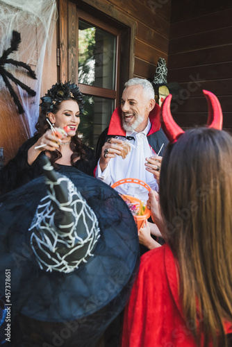 happy couple in creepy halloween costumes holding candies and smiling near blurred kids